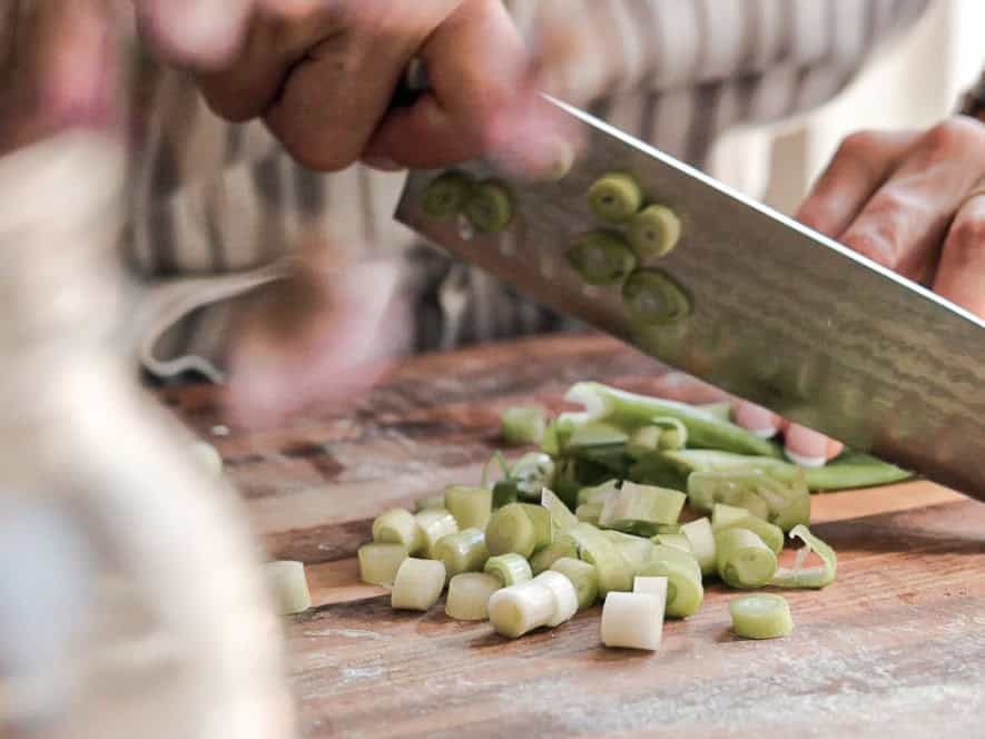 A person chopping green onions on a wooden cutting board. The focus is on the hands and the knife, with sliced green onion pieces scattered around.